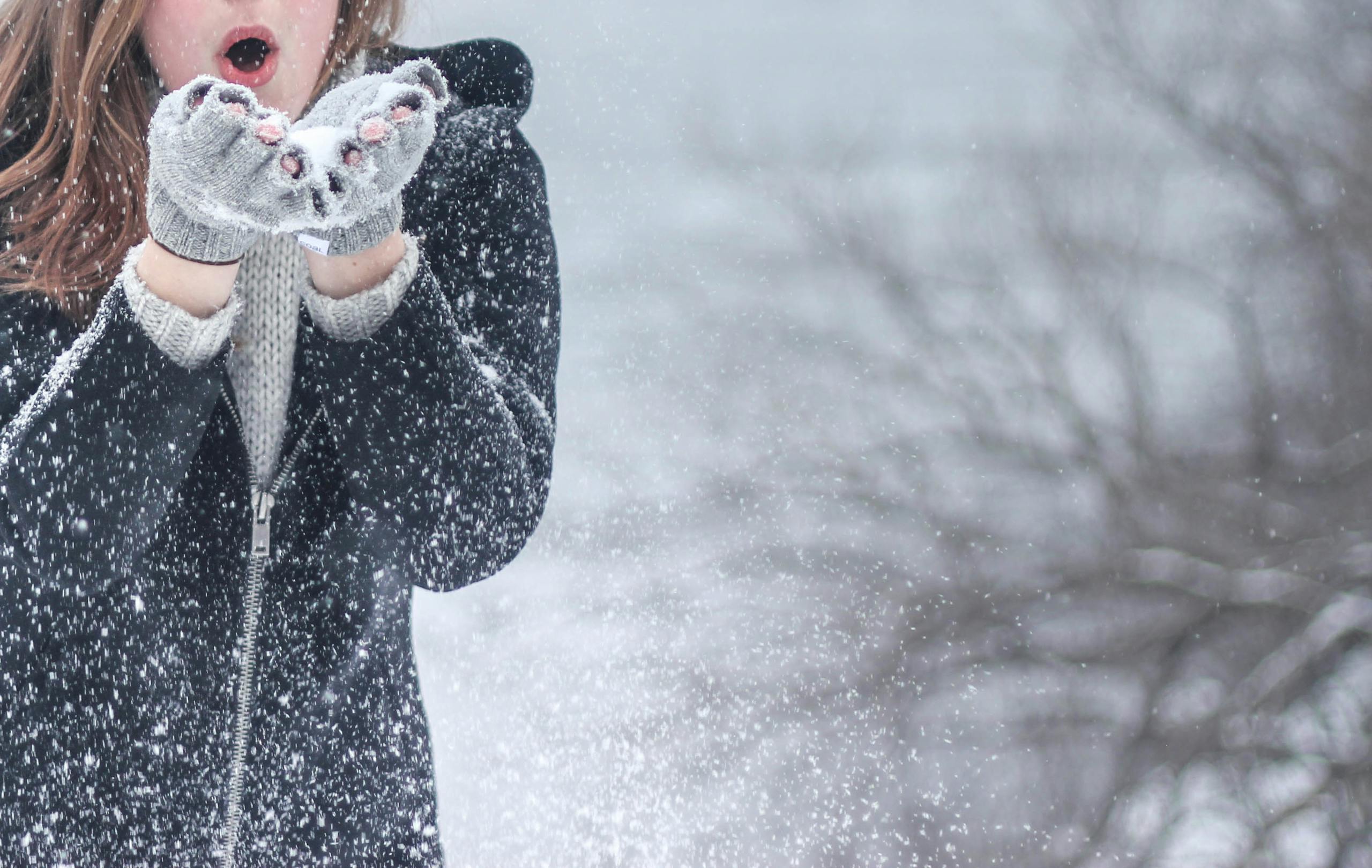 Woman in cozy winter clothing blowing snowflakes with excitement outdoors in a snowy setting.